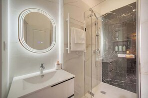 A modern bathroom with a white vanity, a large mirror with lights, a glass shower enclosure, and marble walls. - EMILY 2BR 2baths with AC near the Palais (Cannes)