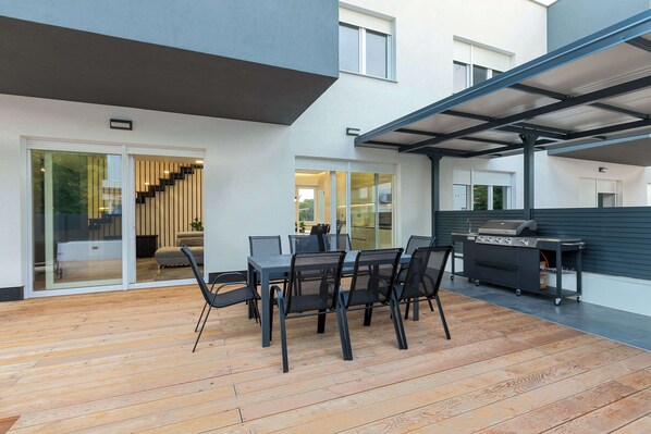 A modern patio with a dining table and chairs, a barbecue grill, and a covered area with a ceiling structure.