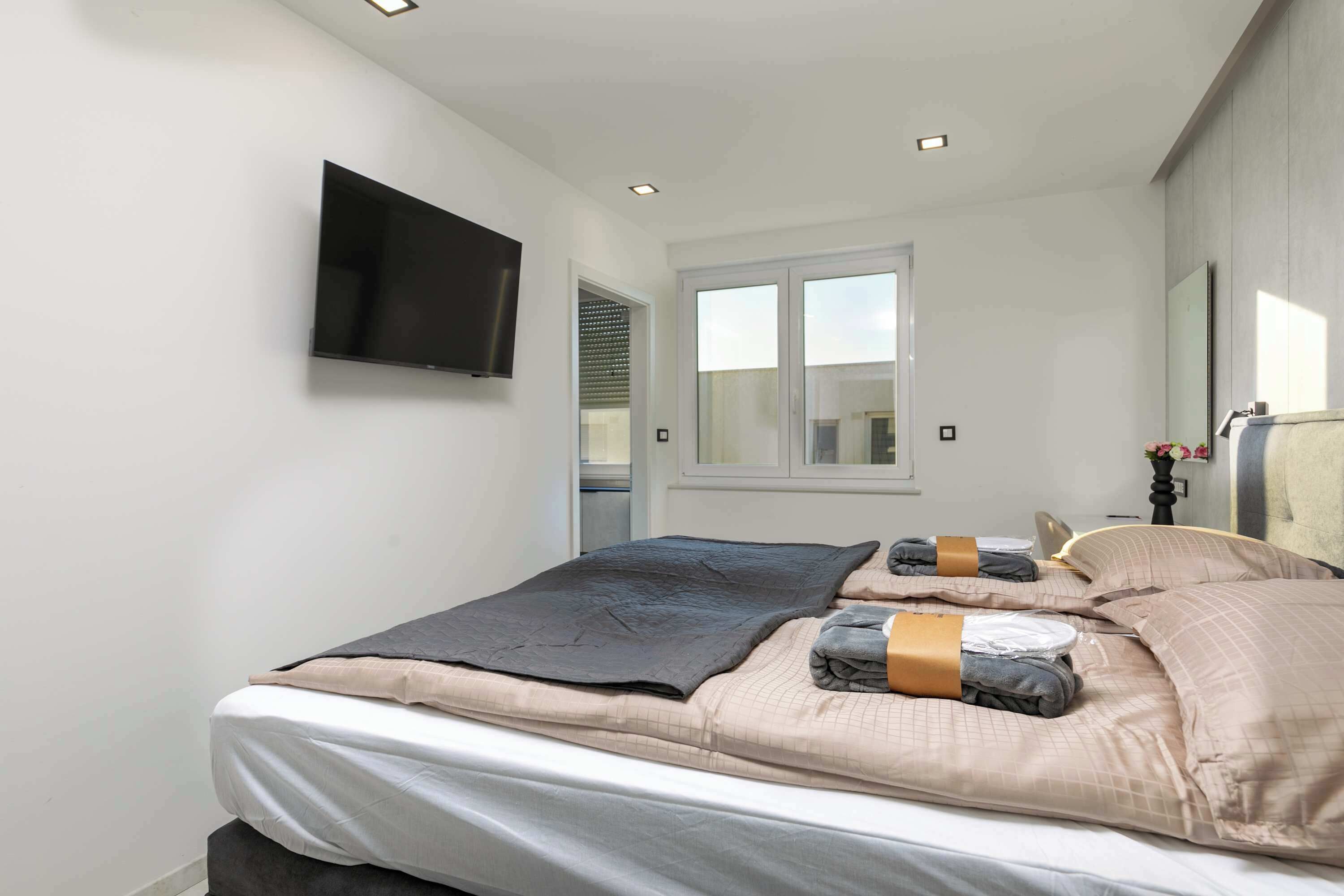 A modern bedroom with a bed, a flat-screen TV mounted on the wall, and a window with blinds.