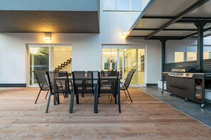 A modern outdoor dining area with a wooden deck, a glass table with chairs, and a barbecue grill.