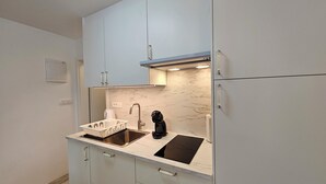 Una cocina compacta con gabinetes blancos, un lavamanos de acero inoxidable, una cafetera negra y un salpicadero de mármol.