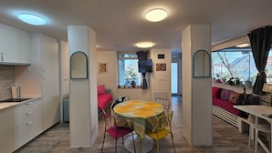 Una cocina moderna con armarios blancos, un comedor con mesa amarilla y sillas rojas, y una sala de estar con sofá y televisor.