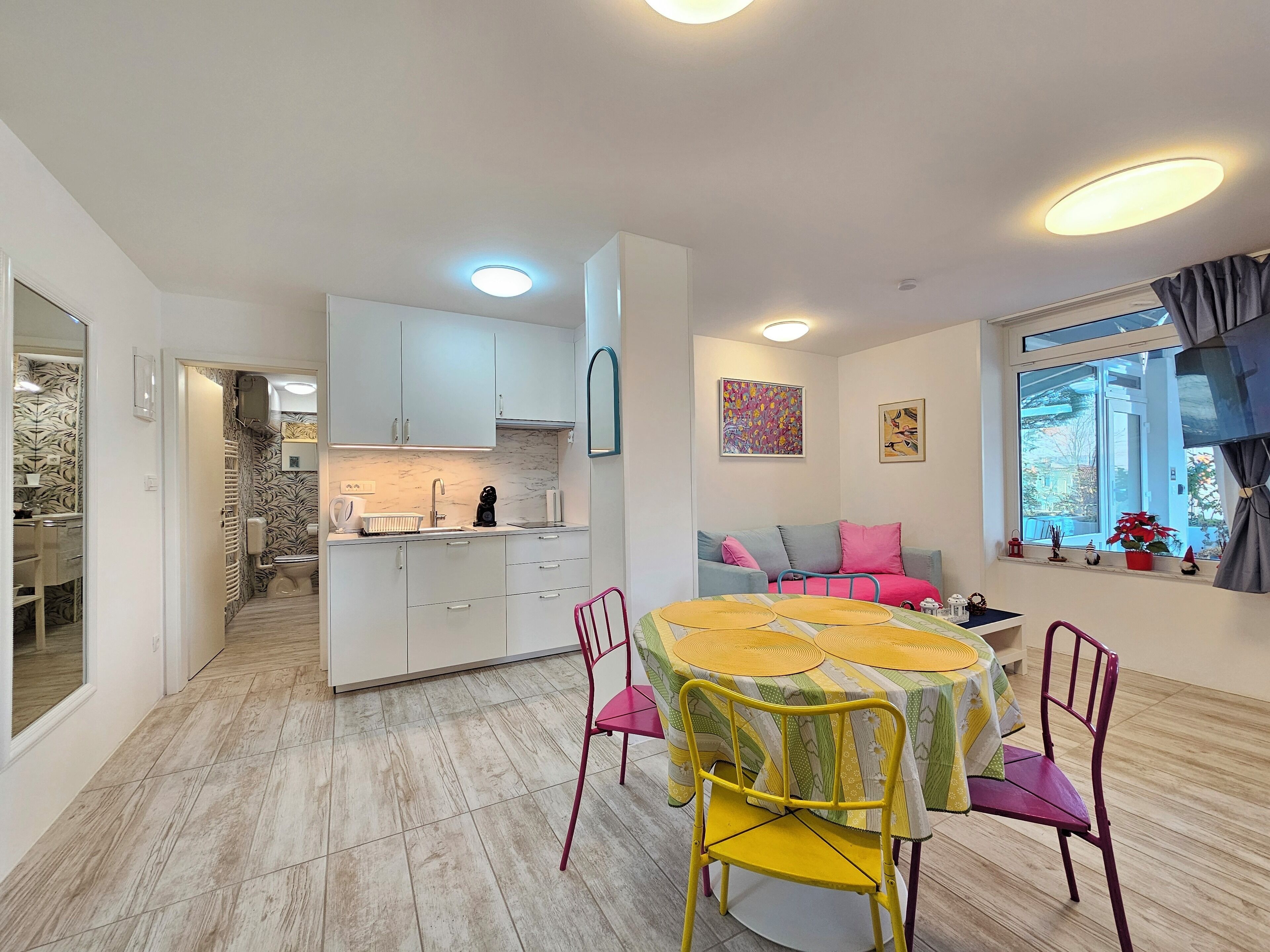 Een moderne keuken met witte kasten, een eethoek met een ronde tafel en kleurrijke stoelen, en een gezellige zithoek met een bank en kussens.