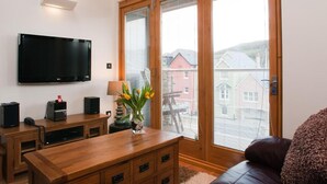 A modern living room with a flat-screen TV, wooden furniture, a glass coffee table with flowers, and a large window with a view of buildings. - Quay Lodge (Dartmouth)