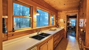 A wooden cabin interior with a kitchen featuring a sink, cabinets, and a window overlooking a forest. - Pines of Three Lakes (Eagle River)