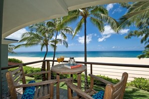 Terraza de madera con mesa y sillas con vista a una playa con palmeras y el océano.