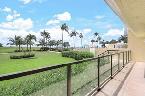A balcony with a glass railing overlooking a well-maintained lawn with palm trees and a clear blue sky.