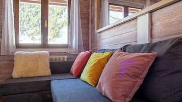 A wooden cabin interior with a sofa and colorful cushions, large windows with curtains, and a view of a forest.