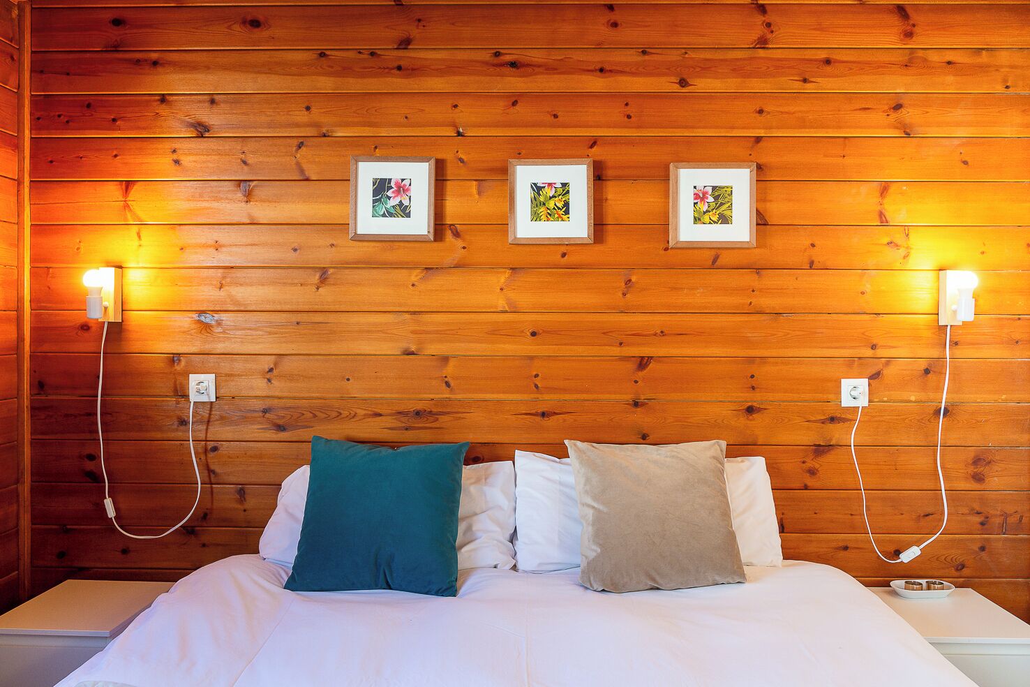 A wooden-paneled room with a bed, three framed pictures on the wall, and two wall-mounted lights.