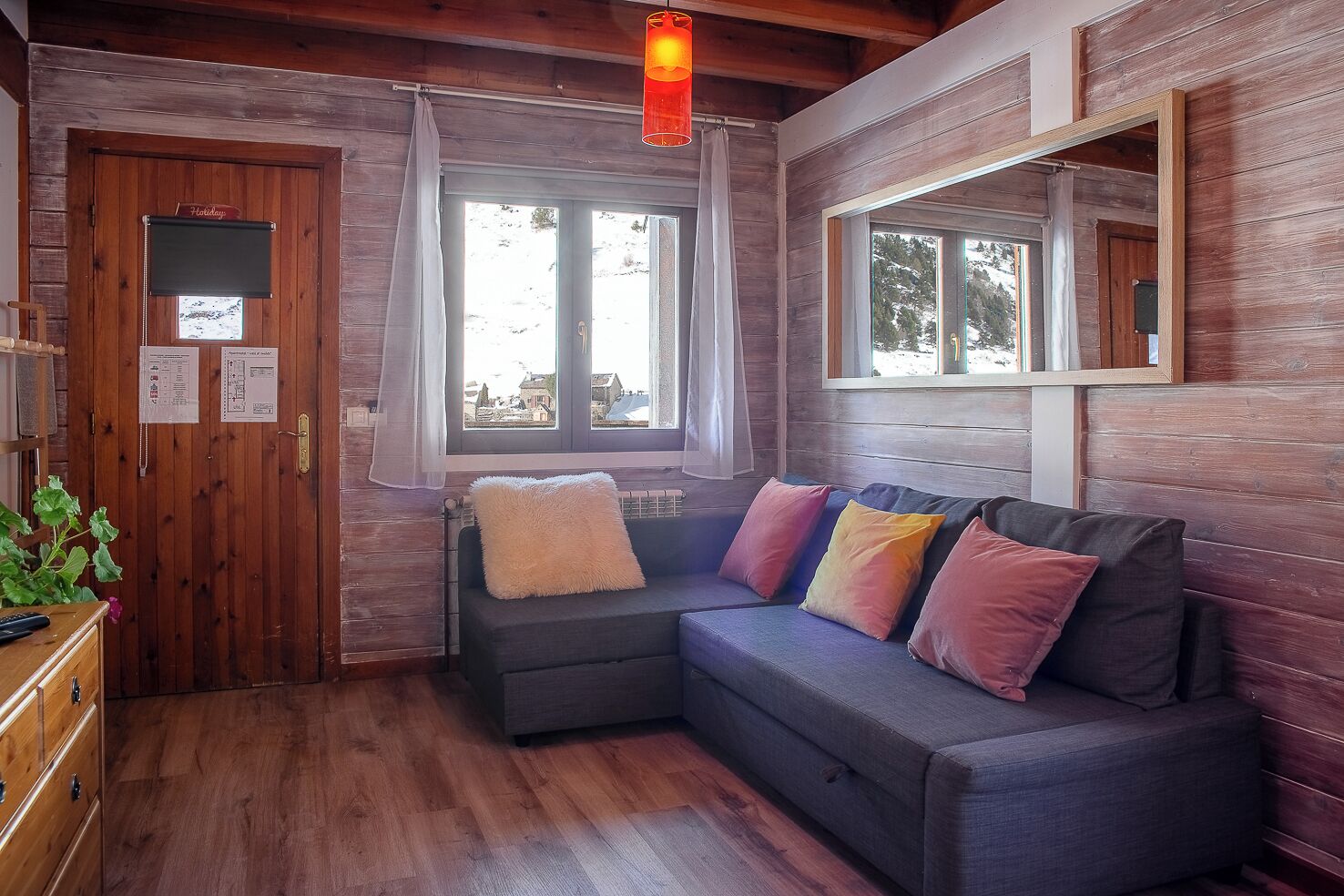 A cozy cabin room with a wooden interior, a sofa with colorful cushions, a window with a view of the outside, and a wooden door with a 'Holiday' sign.