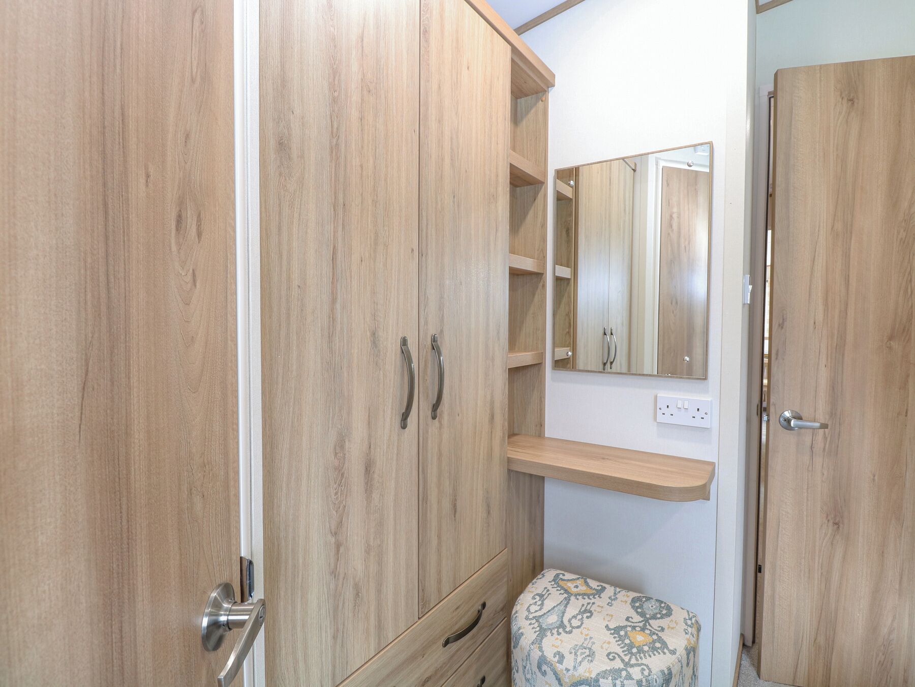 Ein Holzkleiderschrank mit zwei Türen, ein Spiegel darüber und ein kleiner Hocker mit einem gemusterten Sitzkissen.