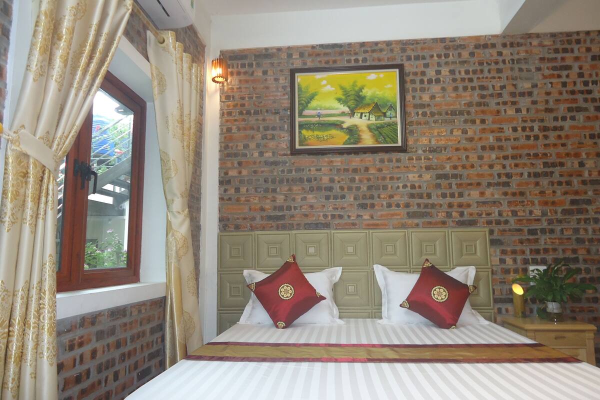 Une chambre d’hôtel avec un mur en briques, un lit avec une literie blanche et des oreillers rouges, un tableau représentant un paysage et une fenêtre avec des rideaux.