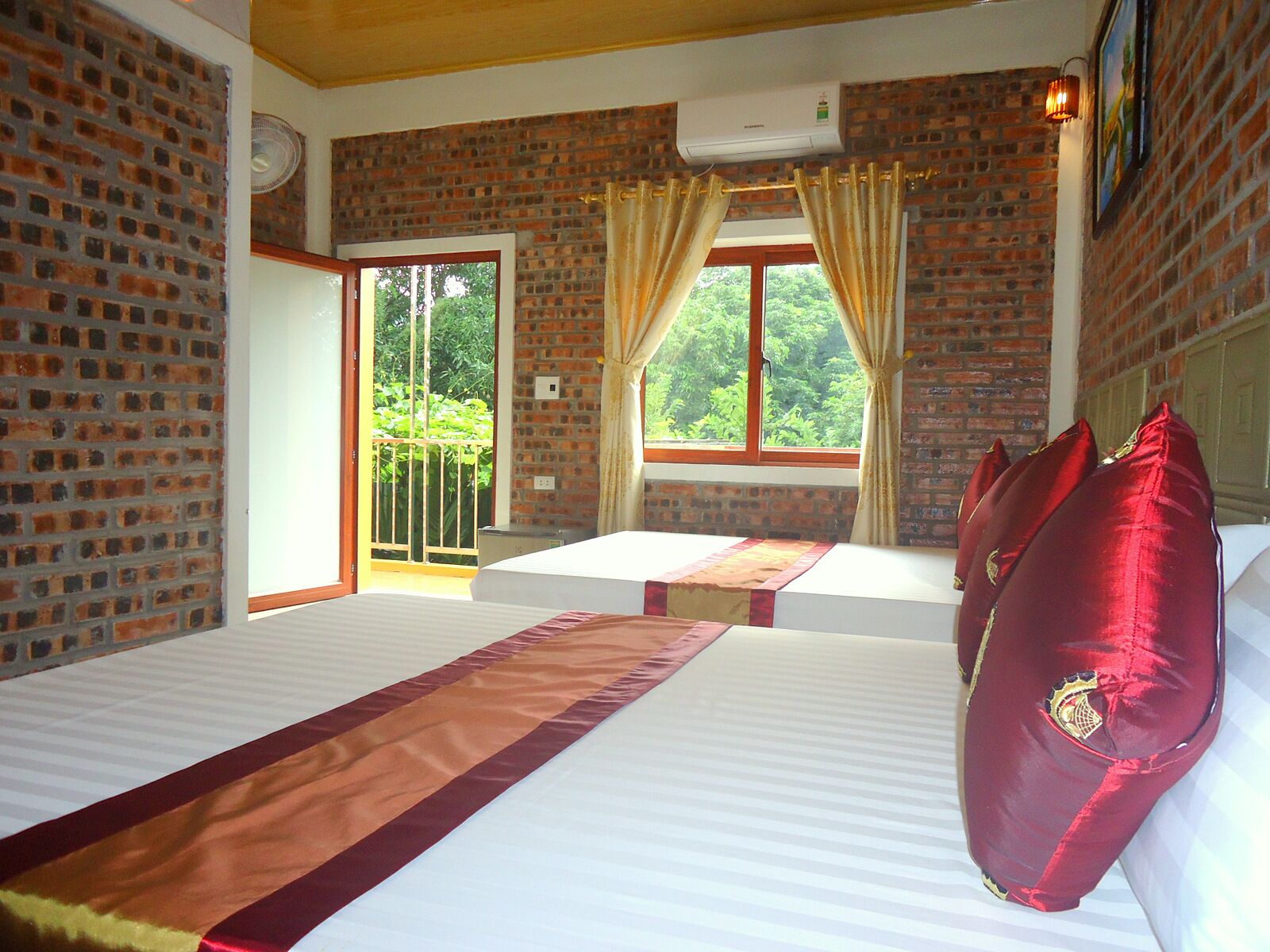Hotelkamer met een bakstenen muur, een groot raam met gordijnen, een bed met een rood kussen en uitzicht op groen buiten.
