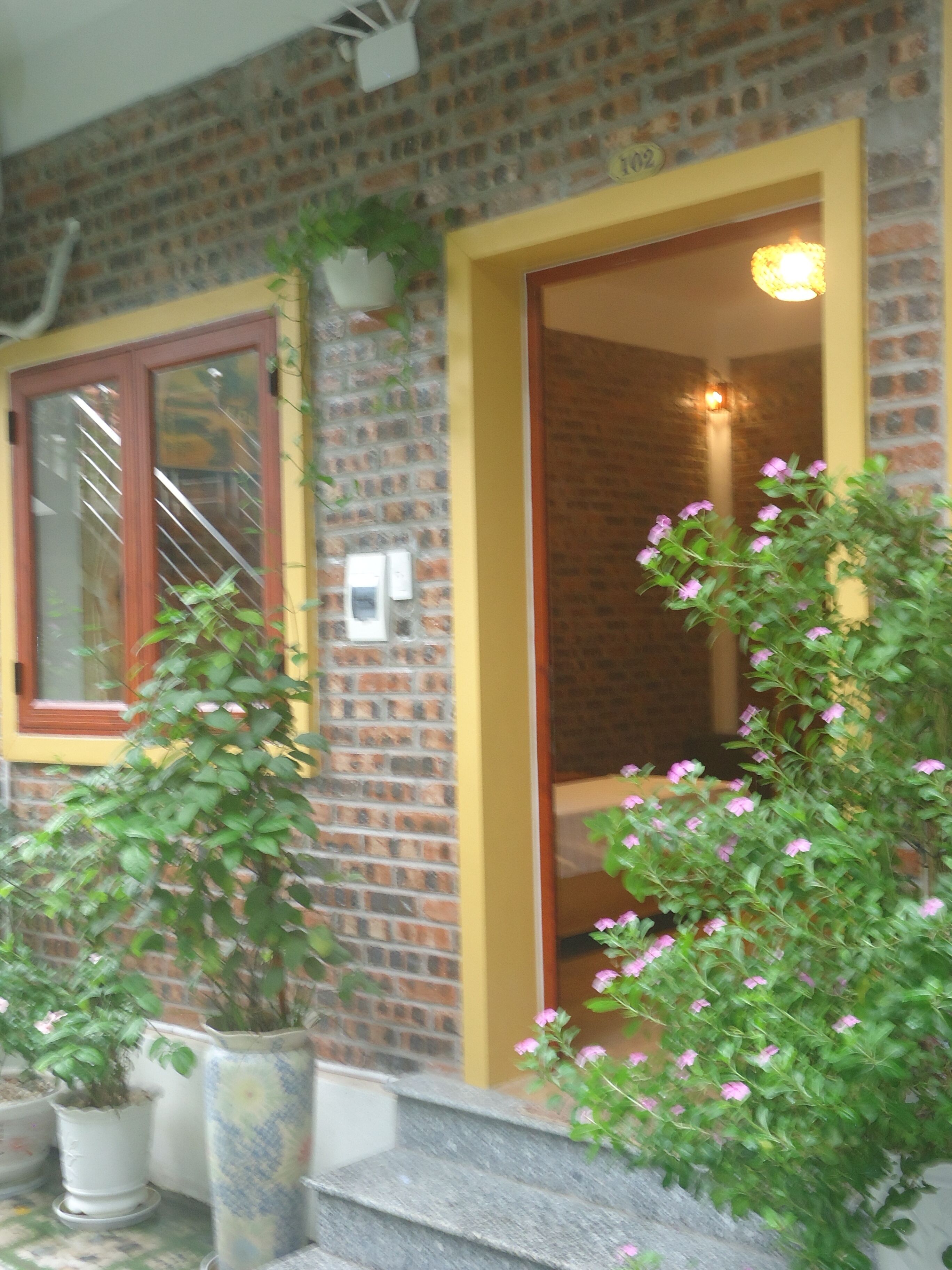 Un bâtiment en briques avec une porte jaune et une fenêtre dotée d’un cadre en bois. On y trouve des plantes en pot et un escalier menant à l’entrée.