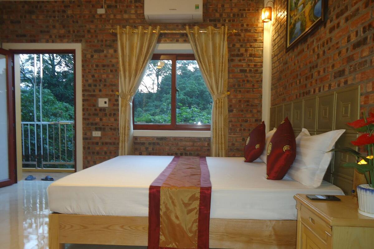 Une chambre avec un grand lit, une table de chevet en bois, une fenêtre avec des rideaux et une vue sur la verdure.