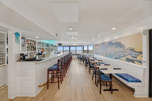 A long dining area with high tables and chairs, a bar on the left, and a large window with a cityscape view.