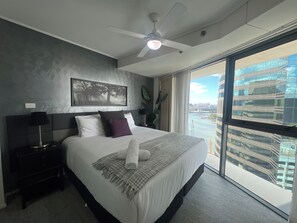 Een moderne hotelkamer met een groot bed, een plafondventilator, stadszicht door raamhoog plafonds en een lijstwerkkunstwerk aan de muur.