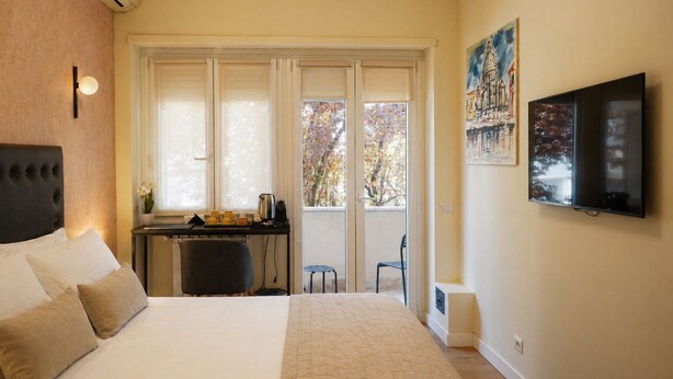 A bedroom with a bed, a desk, a chair, a TV, and a balcony with a view of trees. - Vatican Dolce Remy Guest House (Rome)