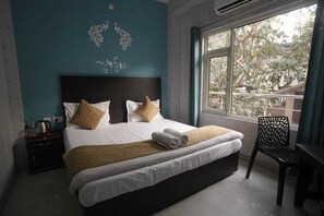 A hotel room with a large bed, a nightstand, a chair, and a window with a view of trees. - Hotel Ananta Inn (Rishikesh)