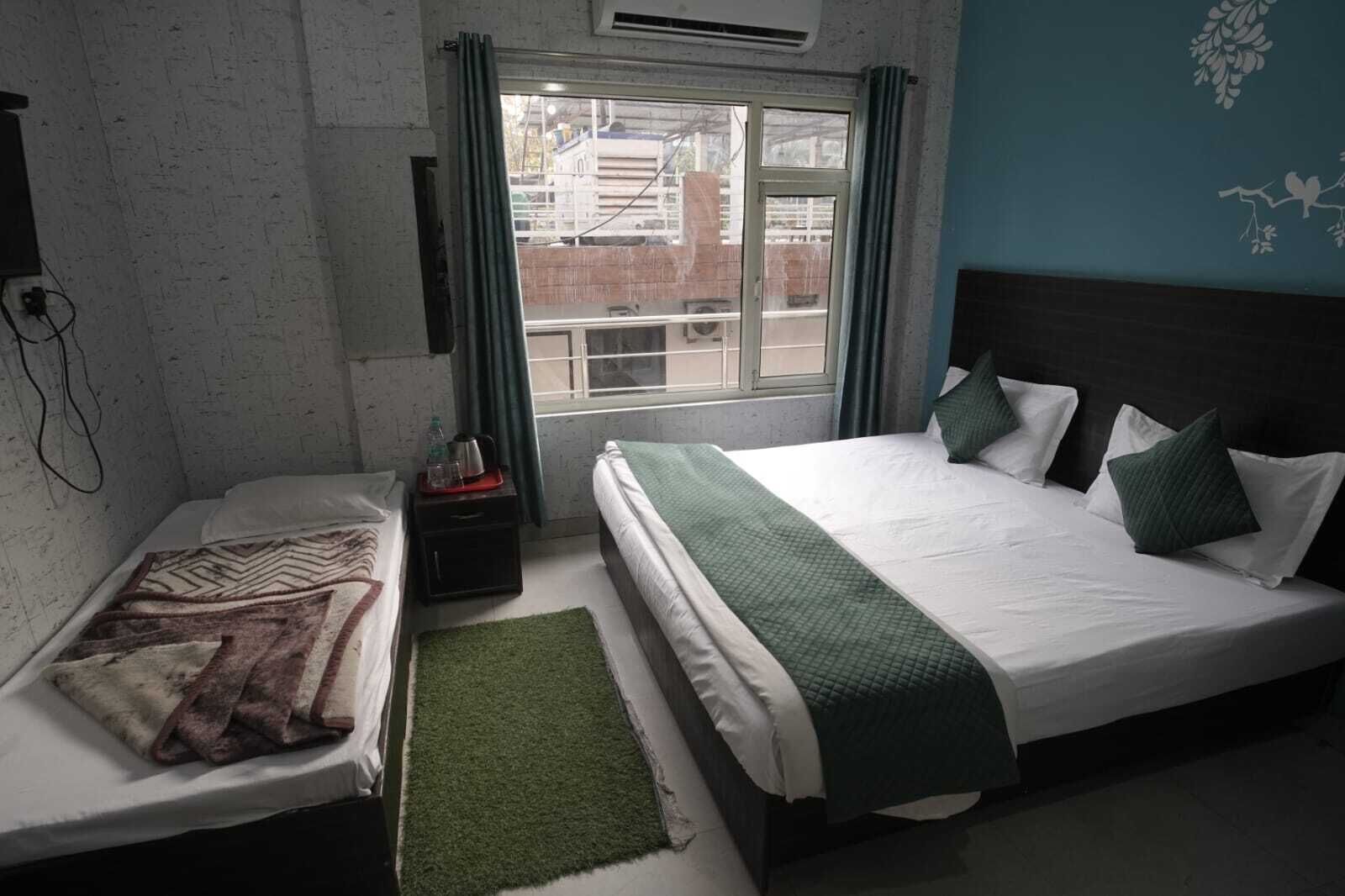 A hotel room with two beds, a window with curtains, a small table with a kettle, and a green rug.
