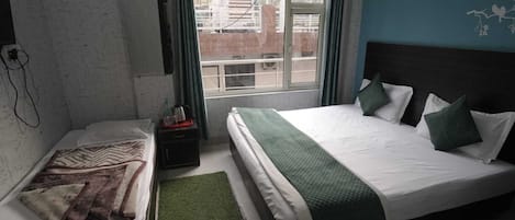 A hotel room with two beds, a window with curtains, a small table with a kettle, and a green rug.