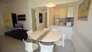 A dining area with a round table and white chairs, a kitchen in the background, and a television on a cabinet. - Villa with private fenced garden in Lignano Pineta (Lignano Sabbiadoro)