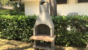 A stone-built outdoor oven with a wooden shelf, surrounded by greenery and a building in the background. - Villa with private fenced garden in Lignano Pineta (Lignano Sabbiadoro)