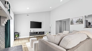 A modern living room with a large sofa, a flat-screen TV mounted on the wall, and a kitchen area in the background. - Quail Ridge 3BR House (Charlotte)