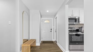 A hallway with a wooden bench, a white door with a stained glass window, and a kitchen with white cabinets and stainless steel appliances. - Quail Ridge 3BR House (Charlotte)