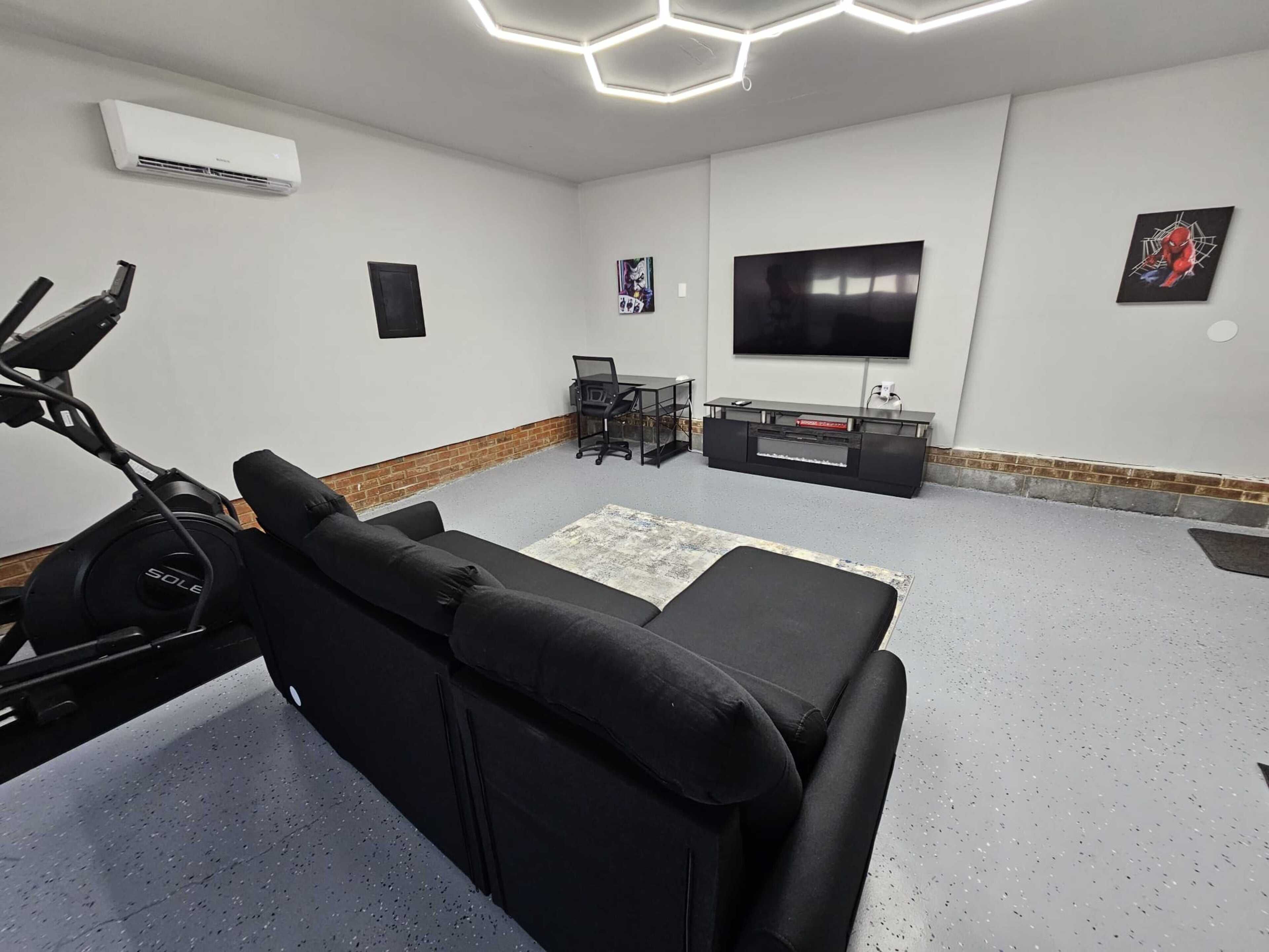 A modern living room with a black sofa, a TV, a treadmill, and a small table with chairs.