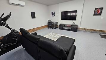 A modern living room with a black sofa, a TV, a treadmill, and a small table with chairs.