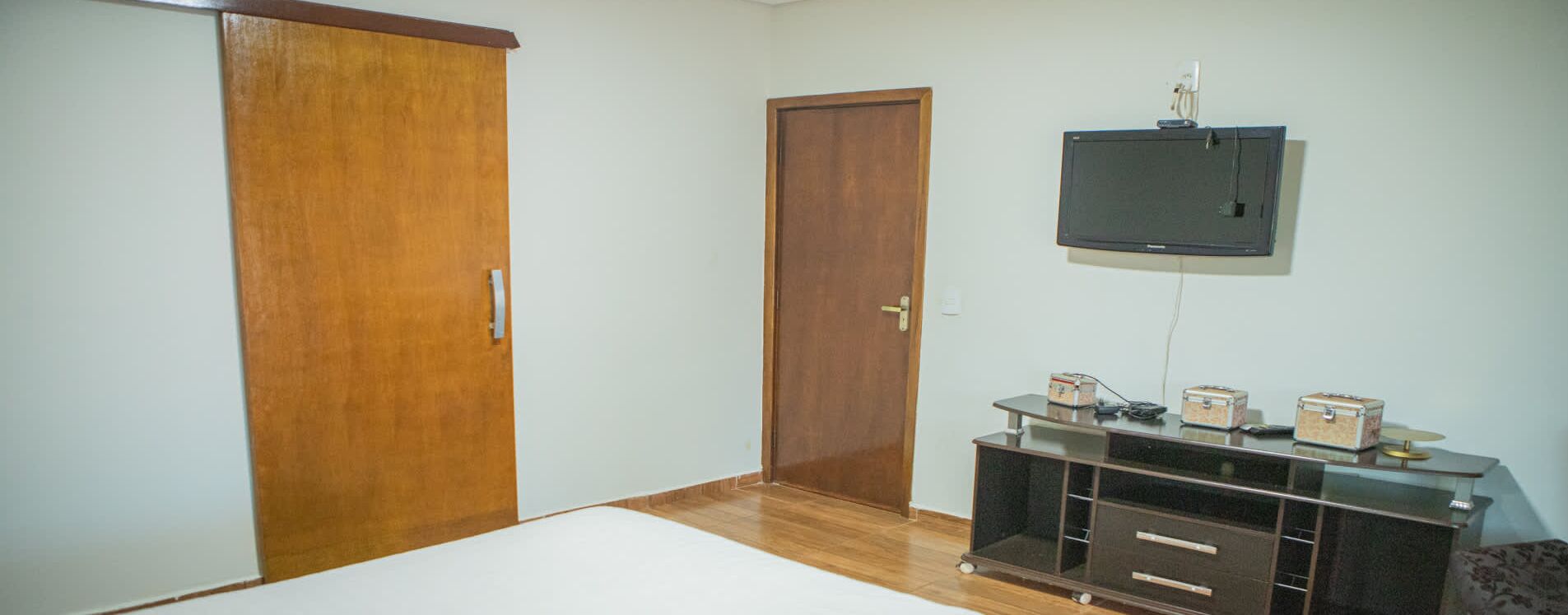 Une pièce comprenant une armoire en bois, une porte fermée, un téléviseur fixé au mur et un petit meuble avec des tiroirs.