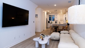 A modern living room with a large flat-screen TV, a sofa, a coffee table, and a dining area with a kitchen in the background. - Modern Designer Stay Bright Chic Spacious (Ontario)