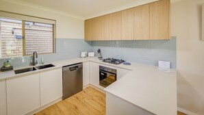 A modern kitchen with white cabinets, a dishwasher, an oven, and a window with a view of a brick wall.