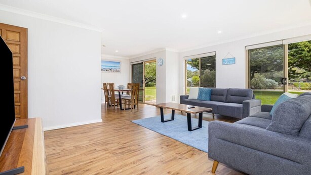A modern living room with a grey sofa, wooden dining table, and chairs. There is a large window offering a view of the outdoors.
