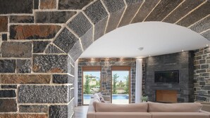 A modern living room with a stone wall, a large arched doorway, a beige sofa, and a flat-screen TV mounted on the wall. - Turtulli Castle Stone Villa 1 - Private Pool Oasis (Hersonissos)