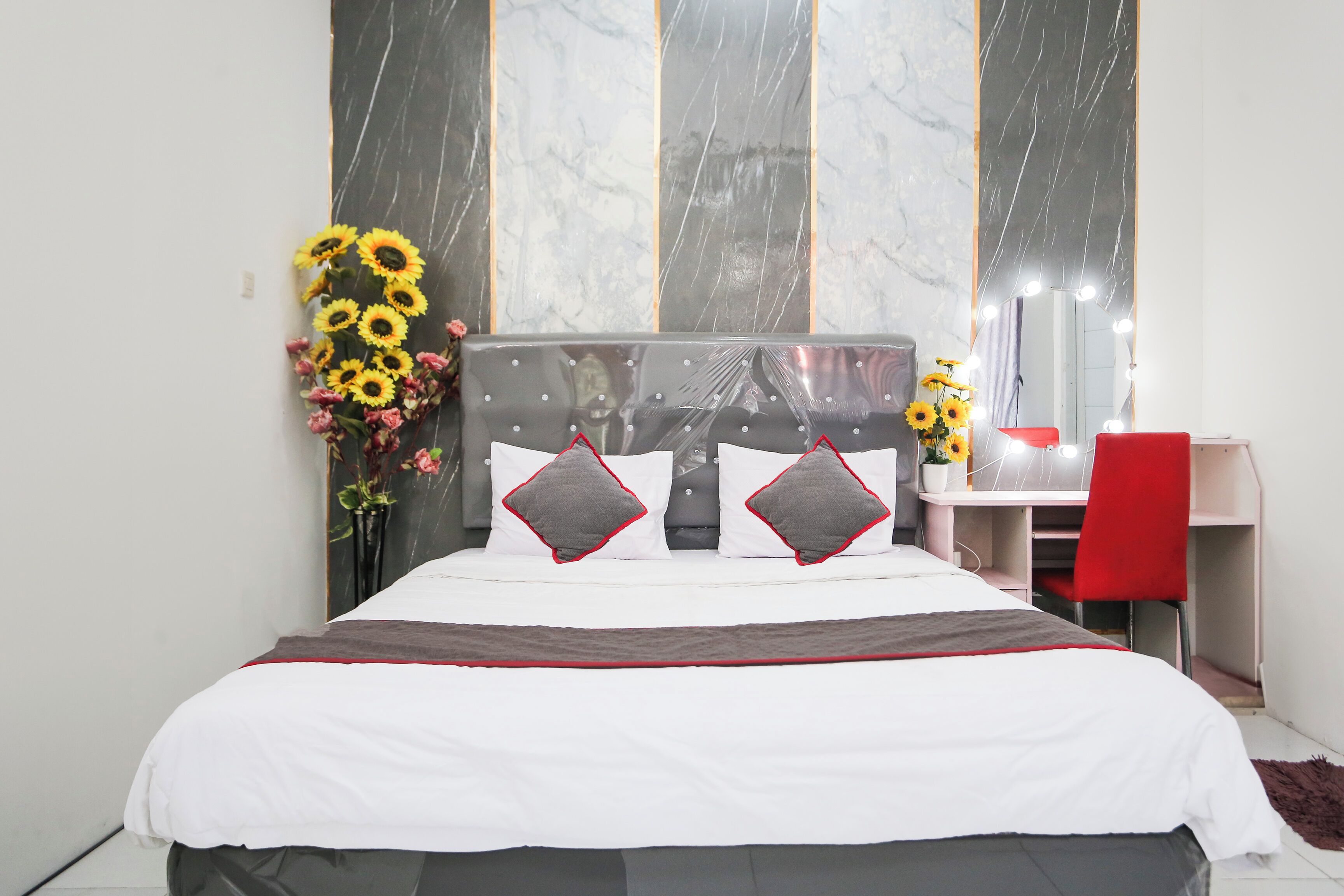 A neatly made bed with a white and gray headboard, a desk with a red chair, and a mirror with lights.