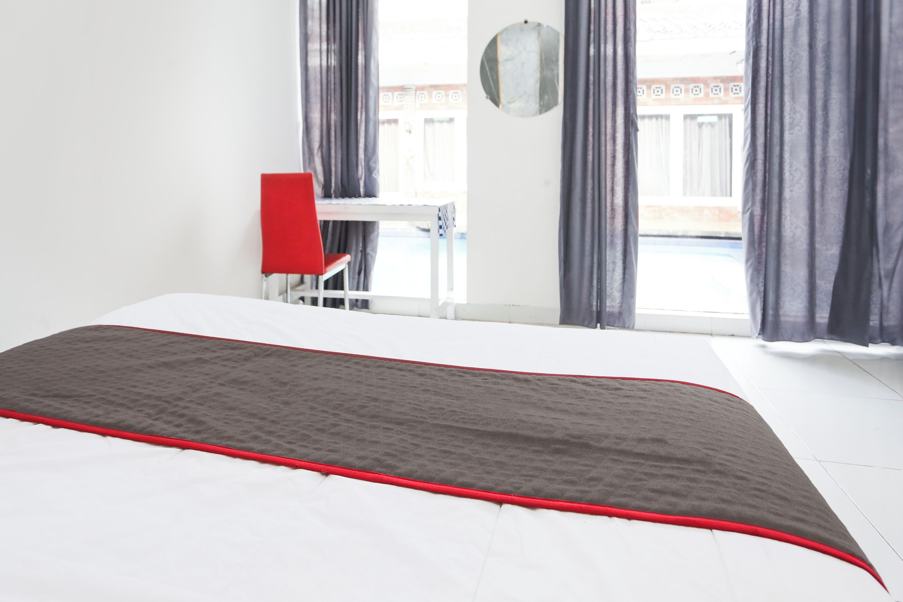 A hotel room with a bed, a red chair, and a view of the outside through large windows.