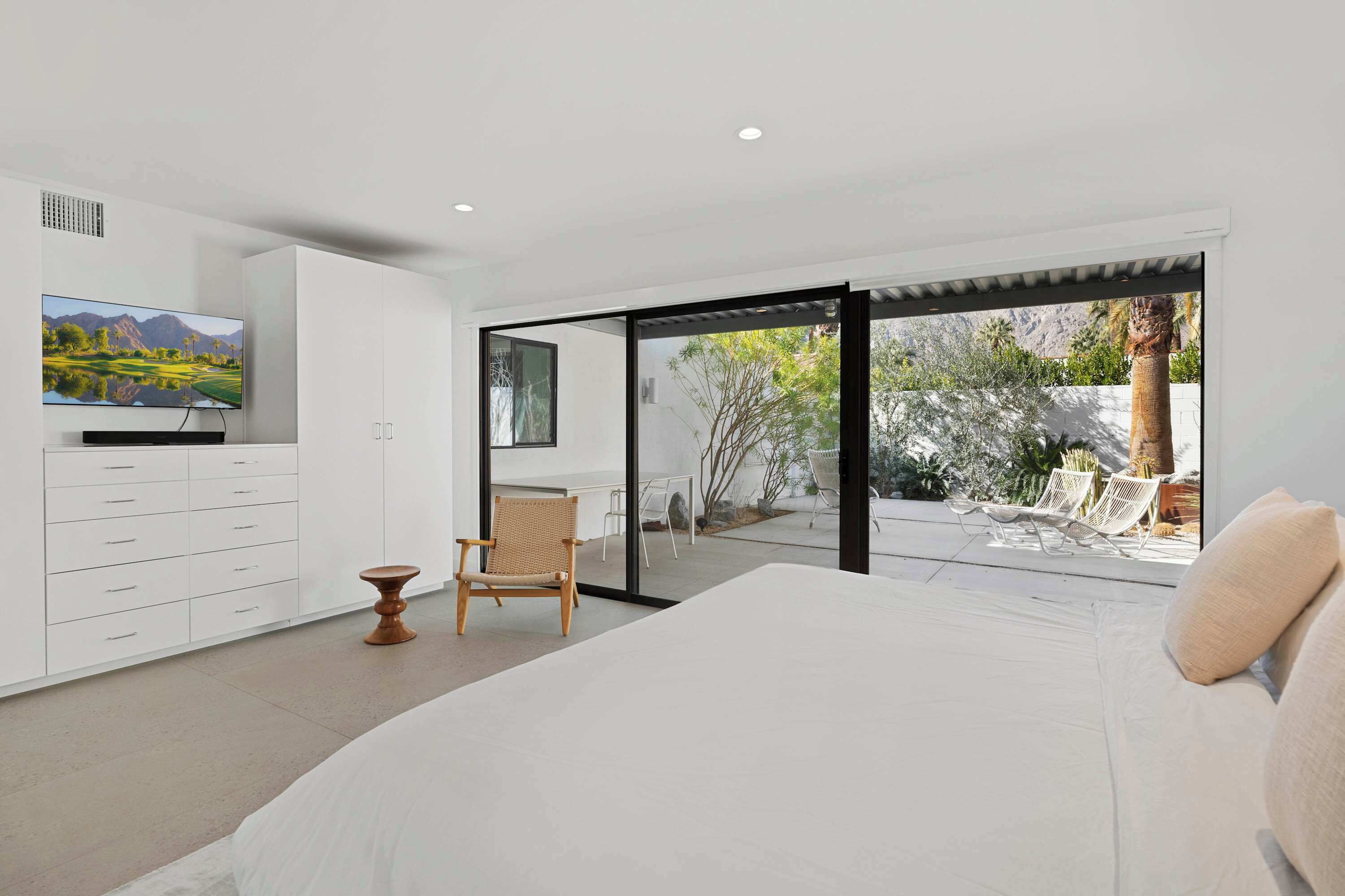 Une chambre moderne avec un grand lit, un téléviseur posé sur un meuble blanc et une porte-fenêtre coulissante donnant sur une terrasse extérieure avec des chaises.
