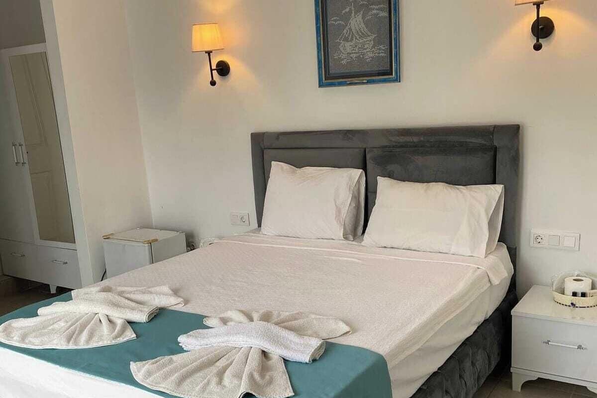 A neatly made bed with a teal bedspread, two white pillows, and a decorative towel arrangement. There are two bedside tables with lamps, a framed artwork above the bed, and a door to the left.