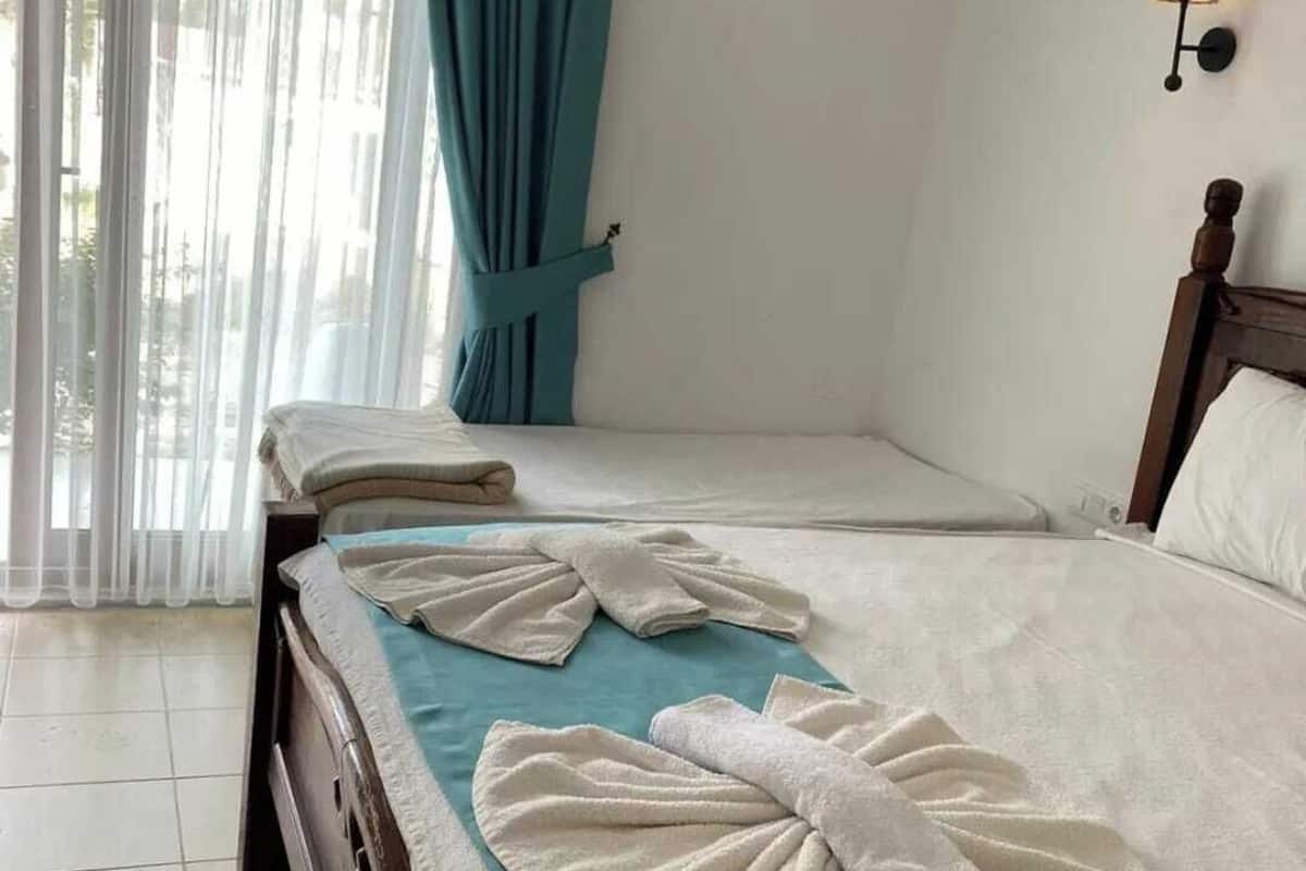 A hotel room with a bed, a bedside table, a lamp, and a view through a window with curtains.