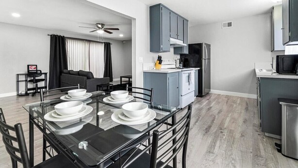 Cocina moderna con armarios grises, un refrigerador de acero inoxidable y un comedor con mesa de vidrio y sillas negras.