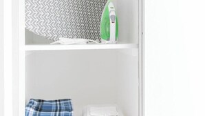A white shelf with a patterned fabric, a green and white iron, folded towels, and a gray laundry basket.