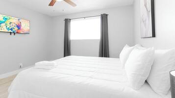 A bedroom with a bed, a ceiling fan, a window with curtains, and artwork on the walls.