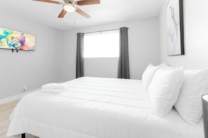 A bedroom with a bed, a ceiling fan, a window with curtains, and artwork on the walls.
