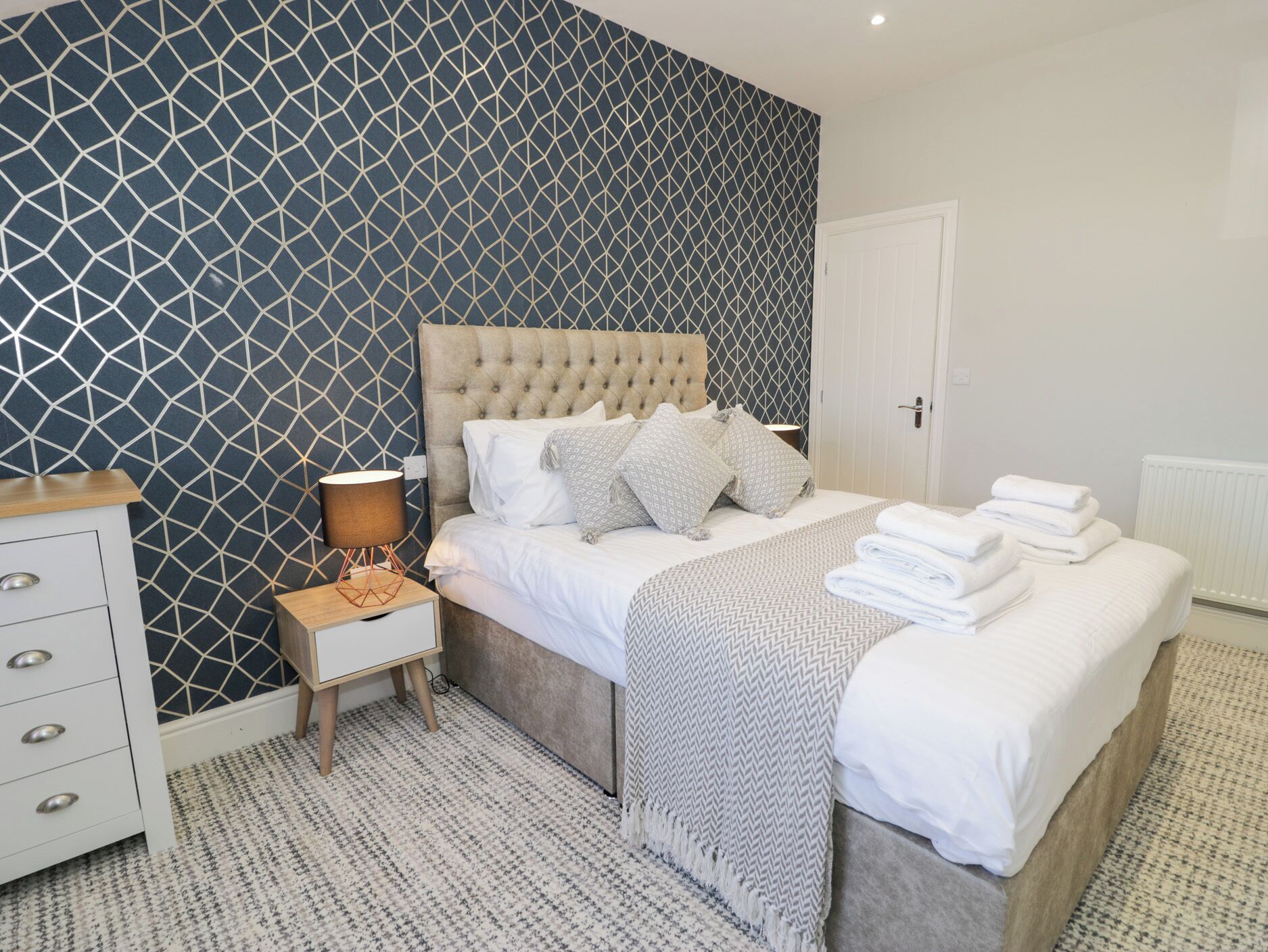 A bedroom with a tufted headboard, a bedside table with a lamp, a dresser, and a neatly made bed with folded towels.