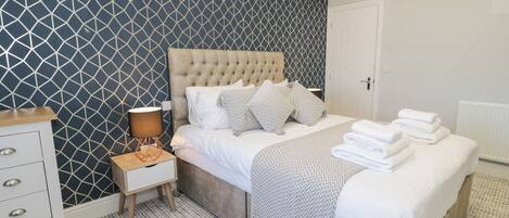 A bedroom with a tufted headboard, a bedside table with a lamp, a dresser, and a neatly made bed with folded towels.