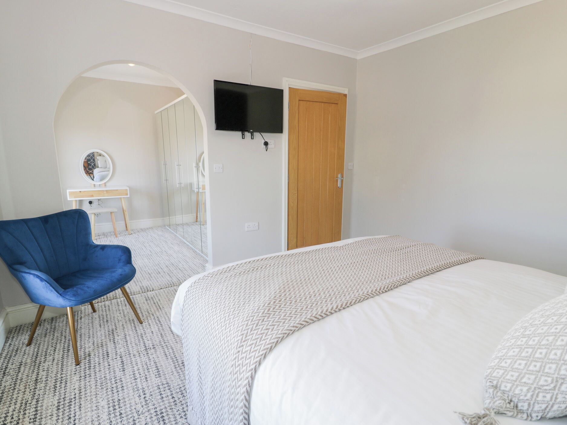A bedroom with a bed, a blue chair, a TV mounted on the wall, and a wooden door.