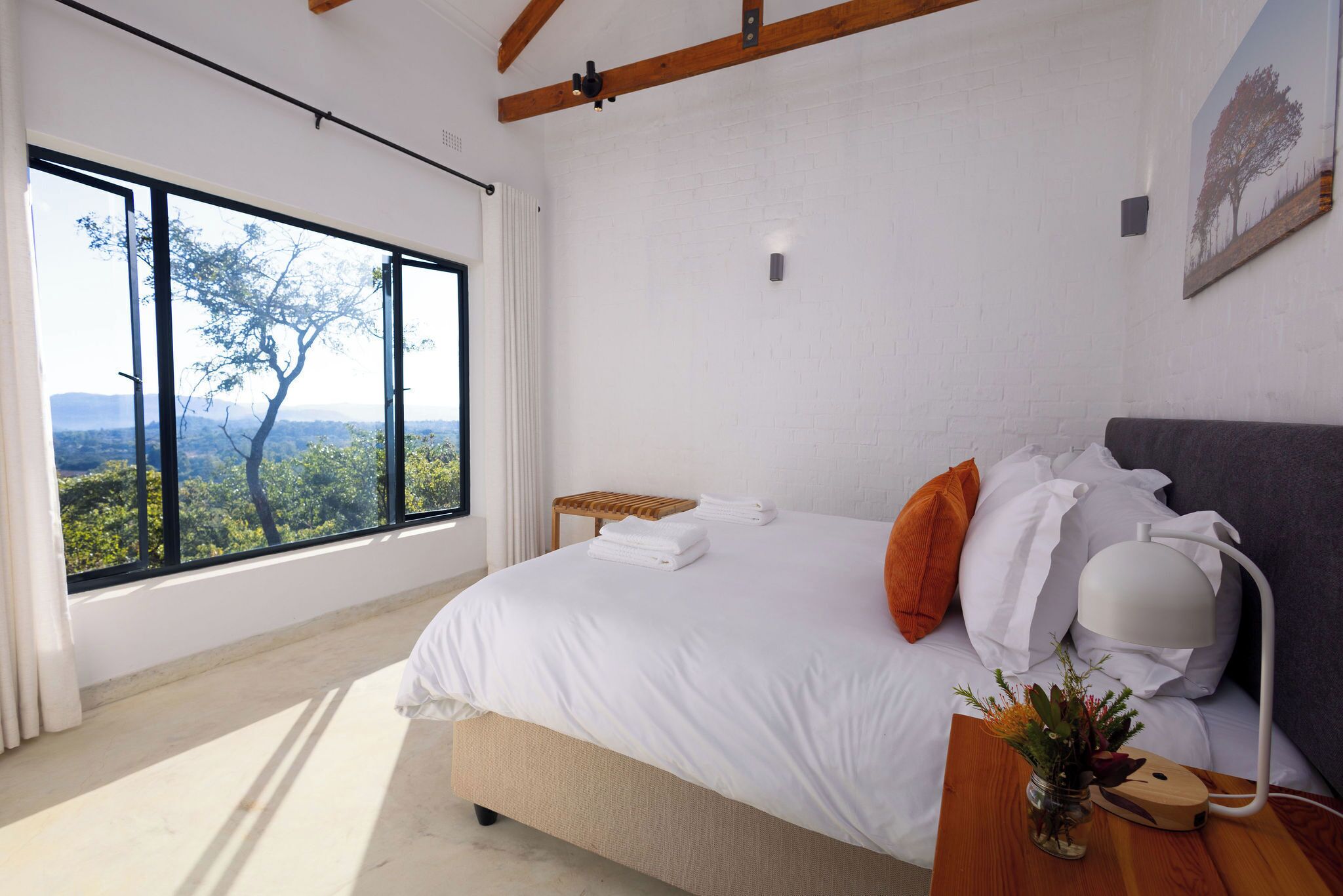 A bedroom with a large window, a bed with white bedding, a bedside table with a lamp, and a view of trees and mountains.