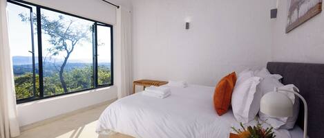 A bedroom with a large window, a bed with white bedding, a bedside table with a lamp, and a view of trees and mountains.